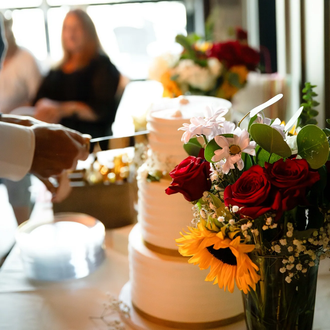A couple cutting their wedding cake beside a bouquet of red roses, daisies and a sunflower