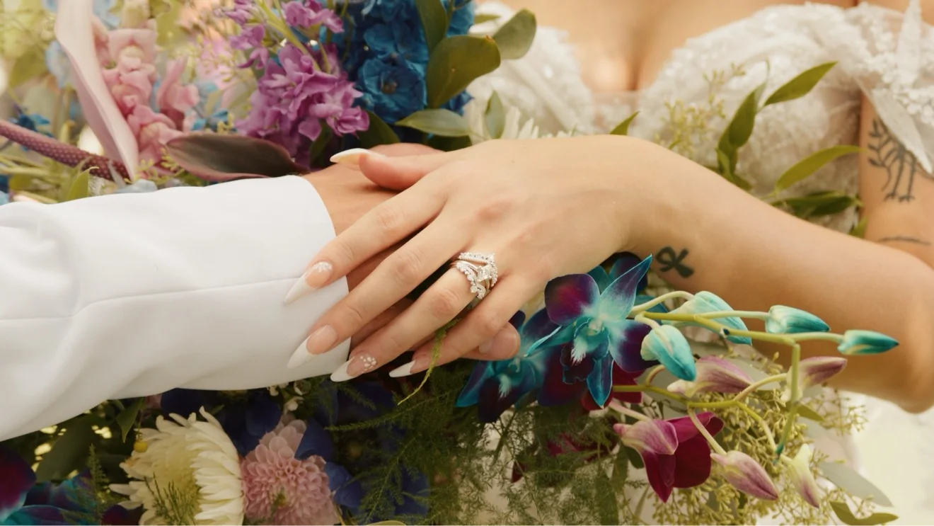 A bride’s hand with her ring resting on the groom’s arm over a bouquet of blue orchids