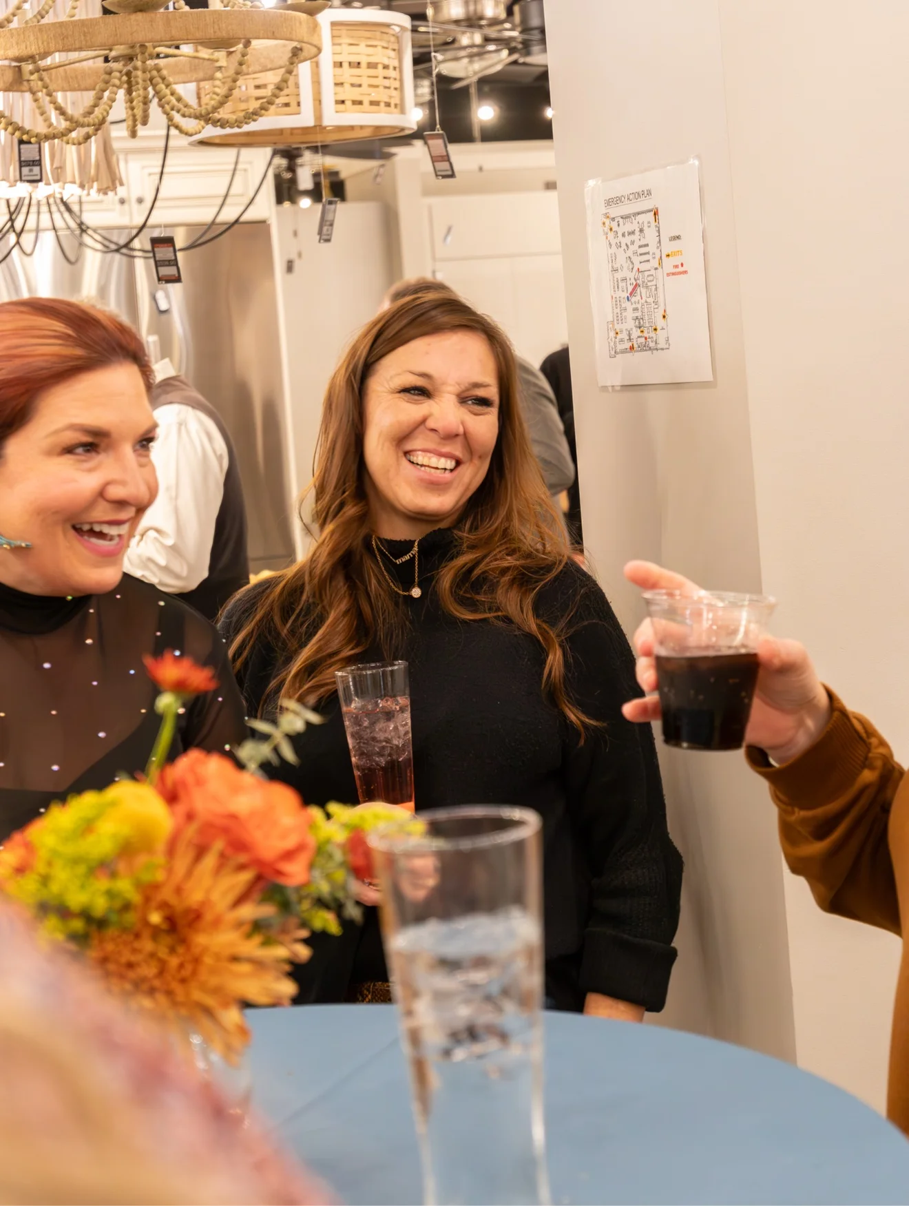 Friends laughing together at an event, drinks in hand, with an orange floral arrangement on the table
